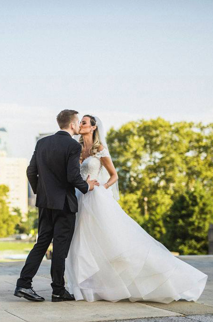 A bride in a cap sleeve wedding dress and veil, and a groom in a suit kiss outdoors, with trees and buildings in the background.