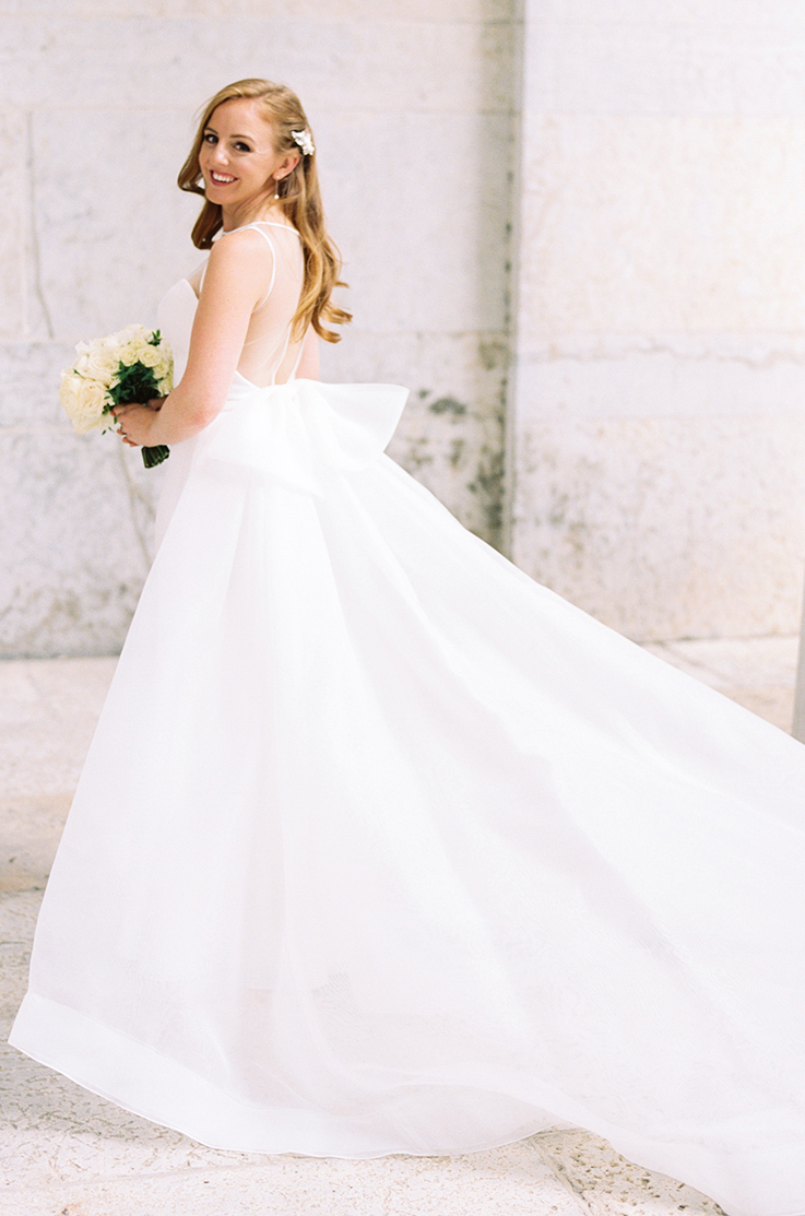 A bride in an Organza skirt wedding dress holds a bouquet while looking over her shoulder and smiling, standing outdoors in front of a concrete wall.