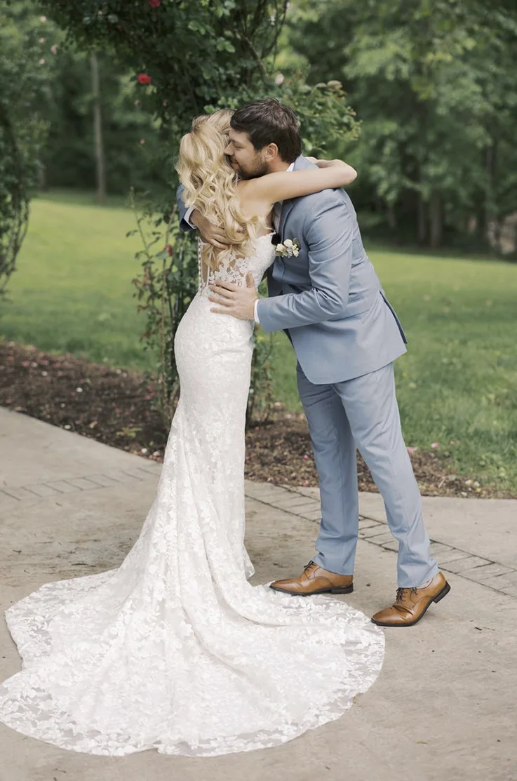 Bride in a lace fit and flare gown with a sheer tulle back with buttons, embraces groom in a blue suit outdoors.