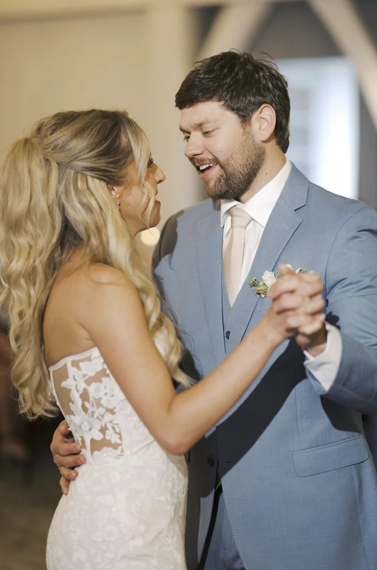 Bride in a strapless lace fitted wedding dress with tulle dances with groom in a blue suit at the reception.