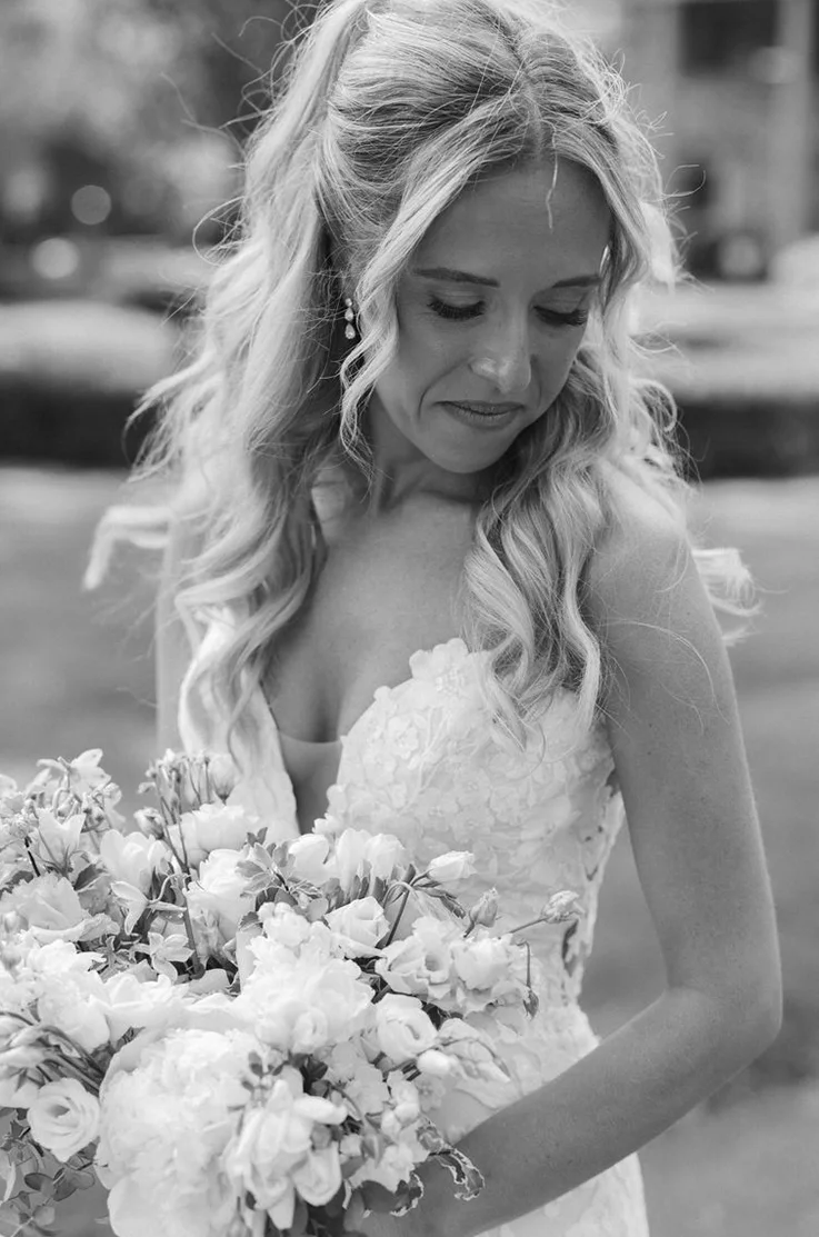 Close-up of bride in a plunging neck lace gown, looking to the side and is holding flowers, in black and white.
