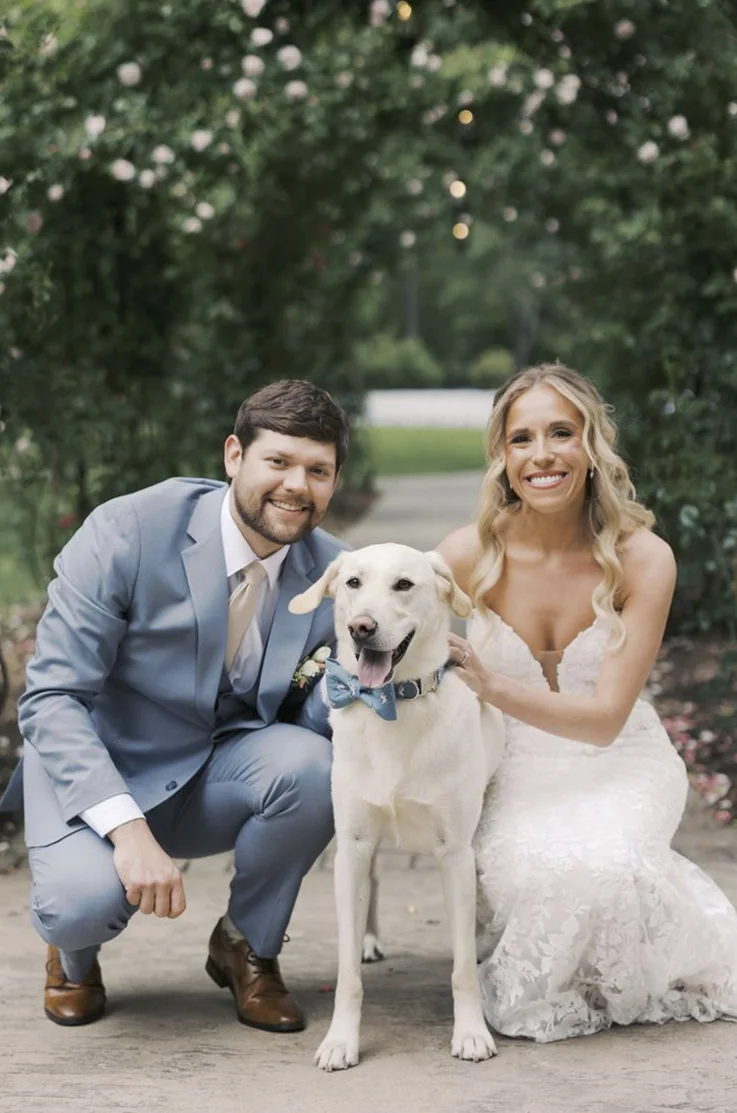 Bride in a strapless lace gown with a plunging neck, bends down and poses with her dog and groom on an outdoor path.