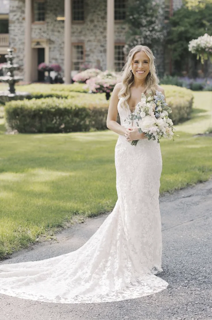 Smiling bride in a fit and flare lace gown with a plunging neck and holding a bouquet on a garden path.