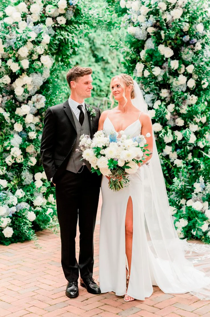 Bride in a plunging neck crepe gown with a skirt slit, is holding a large bouquet and smiling at her groom at lush gardens.