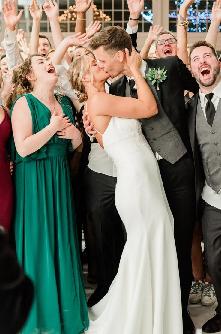 Bride in a low back crepe wedding dress with fit and flare skirt and buttons, kisses her groom as guests cheer.