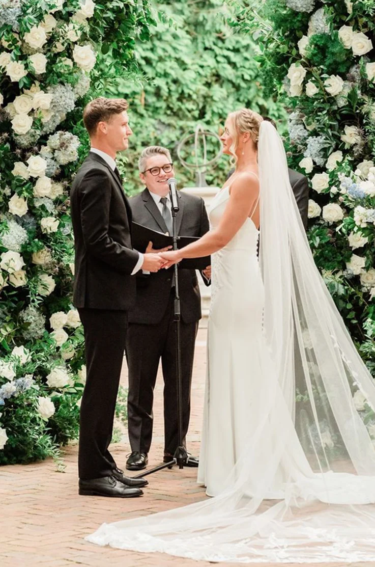 Bride in a fit and flare crepe gown with a buttoned back, and a veil, is facing her groom at the altar between floral arches.