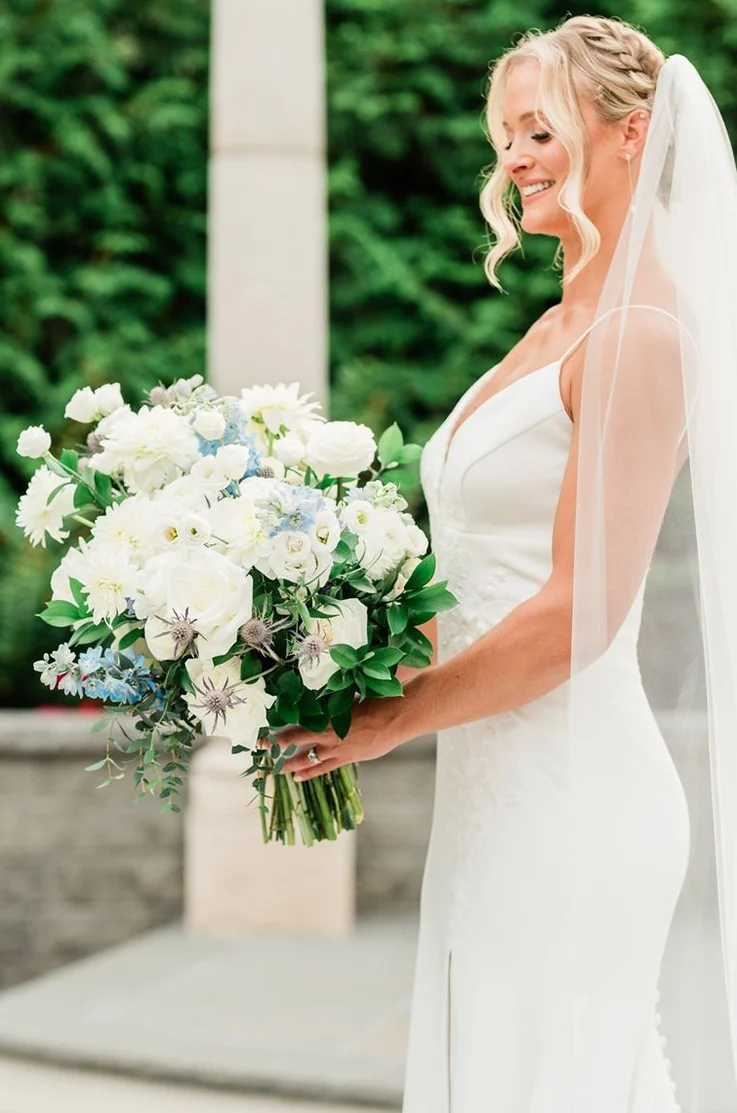 Close-up of bride smiling in a plunging neck crepe gown with sequin lace detail, and a veil, holding a white and blue bouquet. 