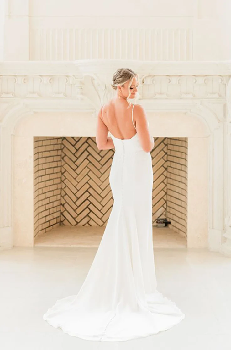 Bride posing in a low back fit and flare gown with buttons and spaghetti straps, standing indoors by a white stone fireplace.