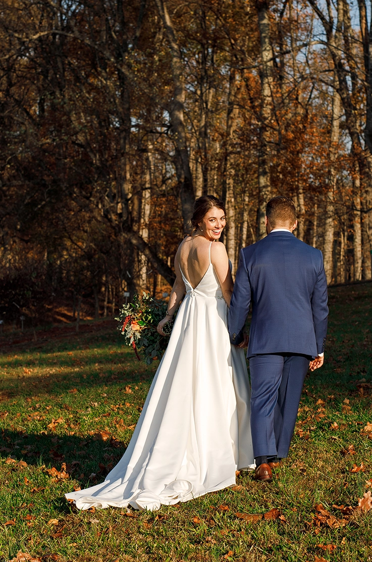 Bride looks over her shoulder in a V-shaped back satin faille gown, walking hand in hand with groom in a sunlit forest.
