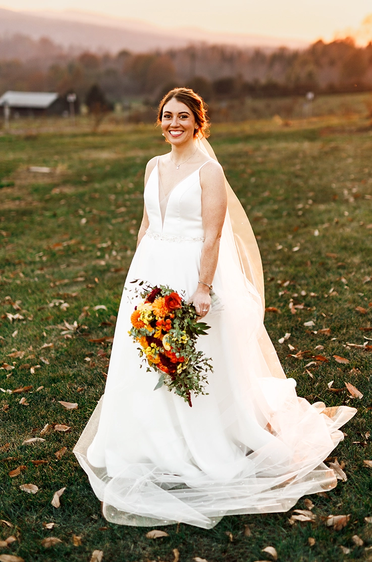 Smiling bride in a plunging neck satin faille gown with a long veil, holding a bouquet in an open field with autumn leaves.