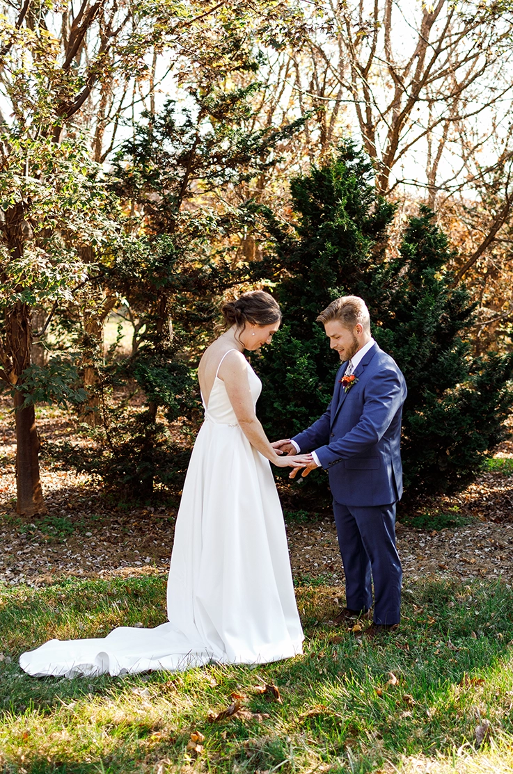 Bride in a deep-V back satin faille gown, faces groom in a blue suit in a sunny forest area. 