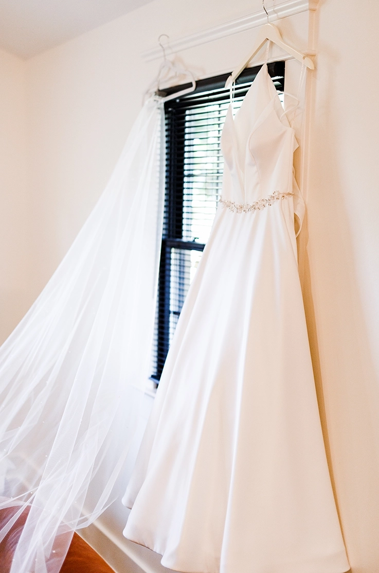 A satin faille A-line wedding gown with a beaded belt and deep plunging neck, hangs on a windows indoors next to a veil. 