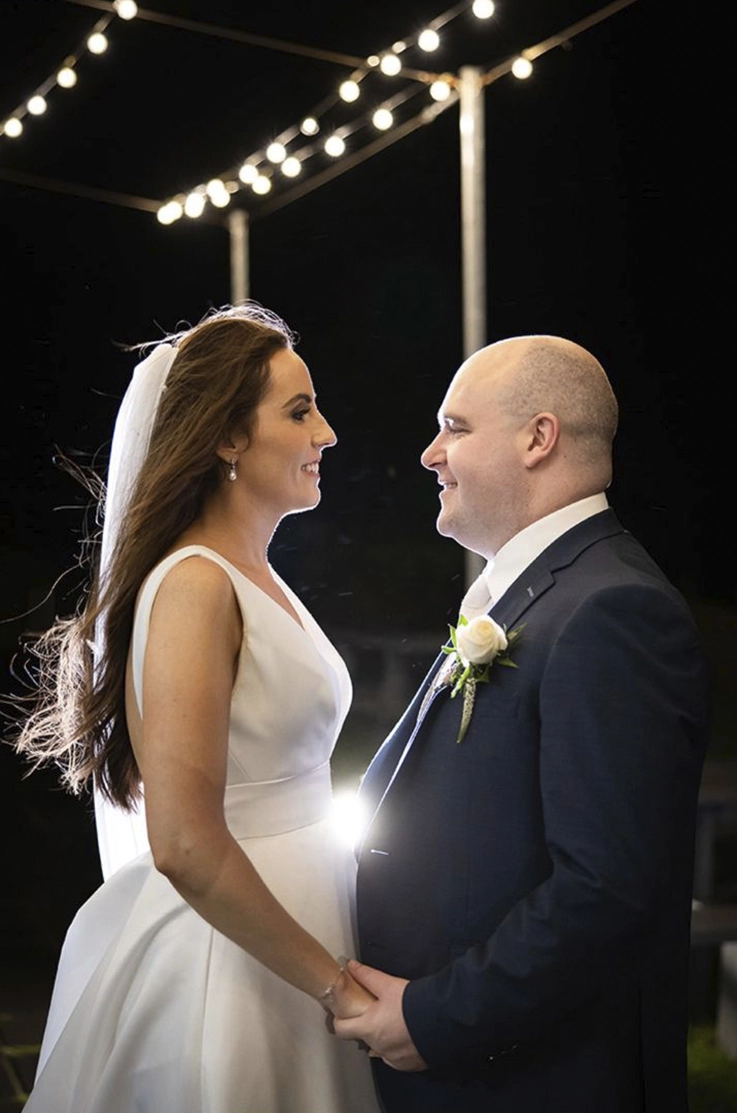 Bride in a sleeveless satin faille gown with a plunging neckline and veil, faces groom under string lights at night.