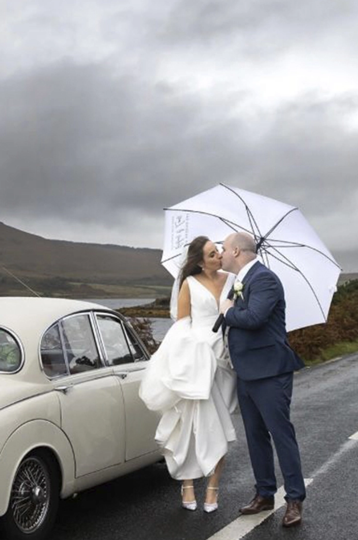 Bride in a draped satin faille wedding dress with a veil, holds her train and kisses groom by a classic vintage car.