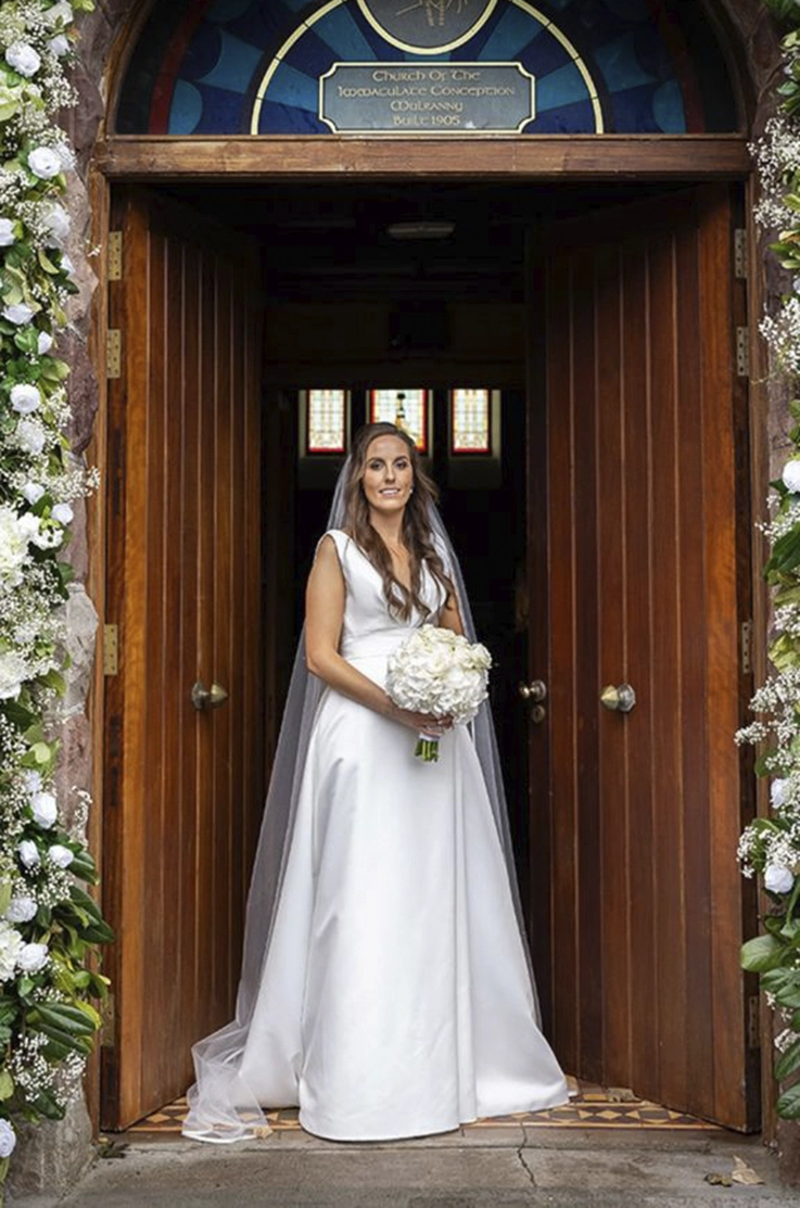 Bride in a draped satin faille bodice gown with a plunging neckline and veil, holds a white bouquet and stands in a doorway.