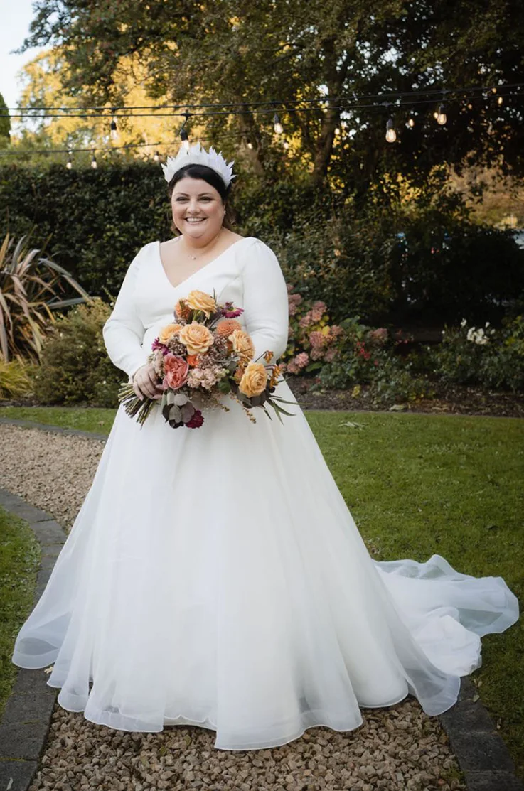 Smiling bride in a crepe bodice wedding dress with a V-neck and organza skirt, holding a bouquet on a garden path.
