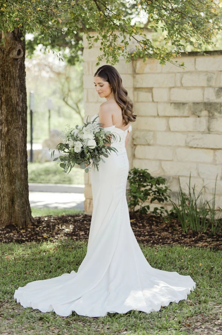 Bride poses in a draped crepe fitted gown with a buttoned back, and sleeves, holding a bouquet by a large tree.