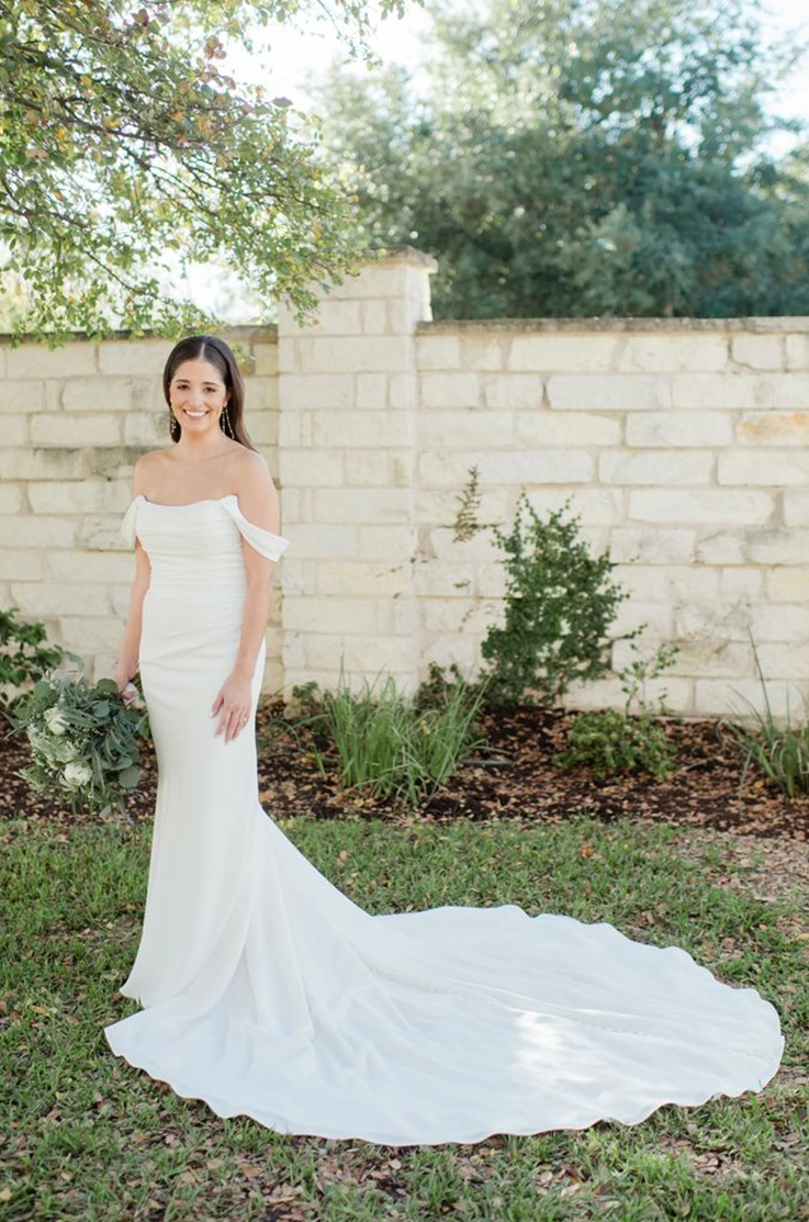 Smiling bride in a draped crepe gown with sleeves and a scoop neckline, posing outdoors by a stone wall.