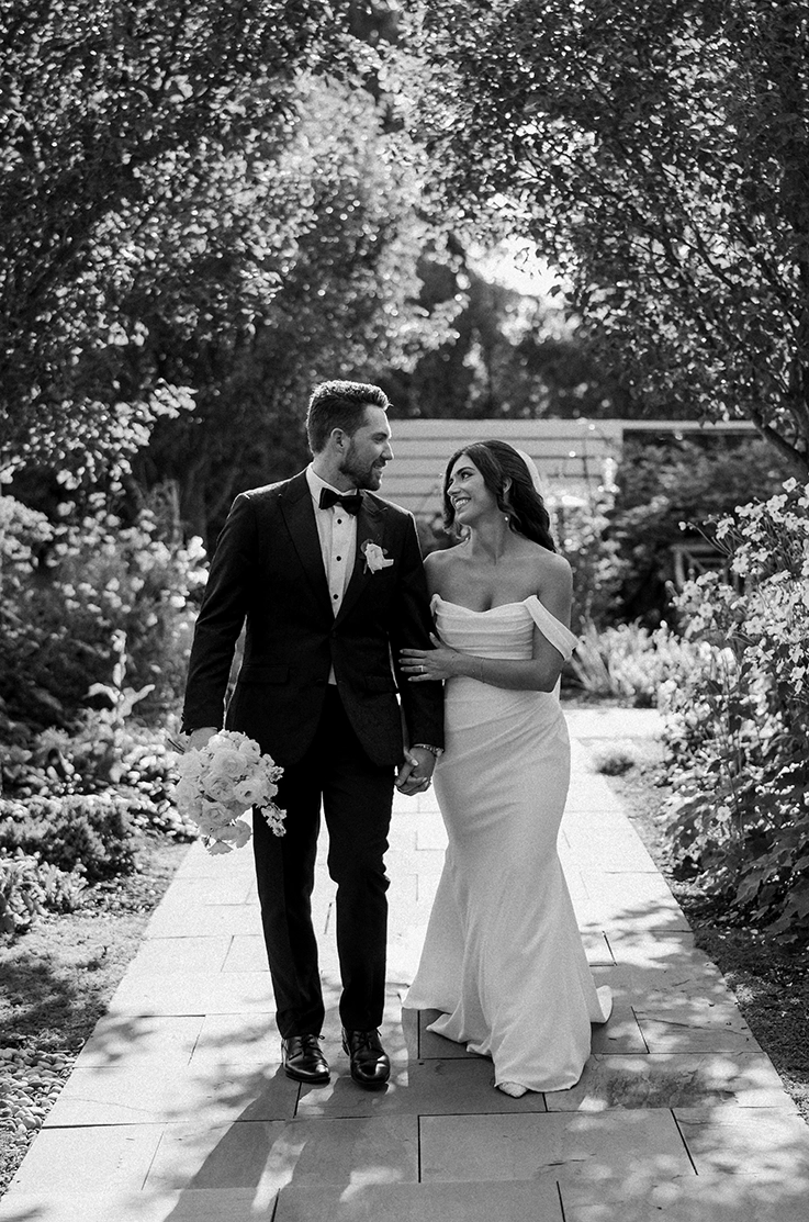 Bride in a draped crepe bodice gown with sleeves, is walking alongside her groom on an outdoor path in black and white.