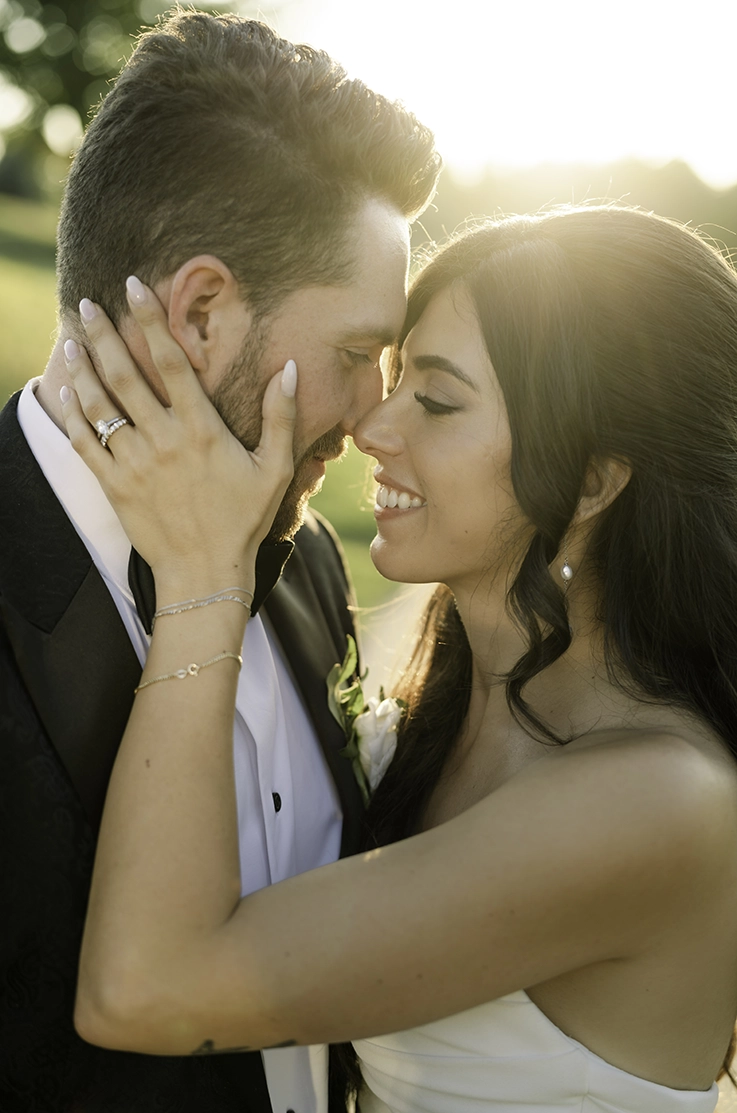 Close-up of bride in a strapless draped crepe gown, embracing her groom in a tuxedo, in the sunlight.