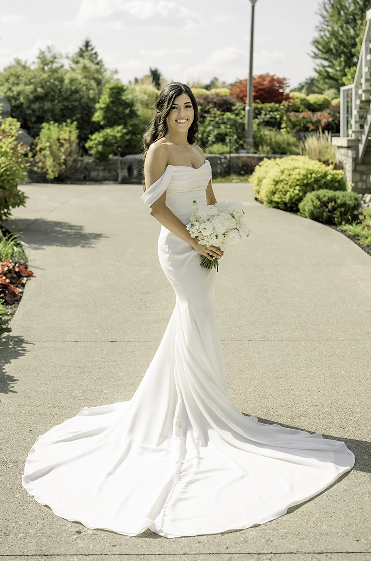 Bride in a crepe draped wedding dress with a scoop neck and off-shoulder sleeves, holding a bouquet and posing outdoors.