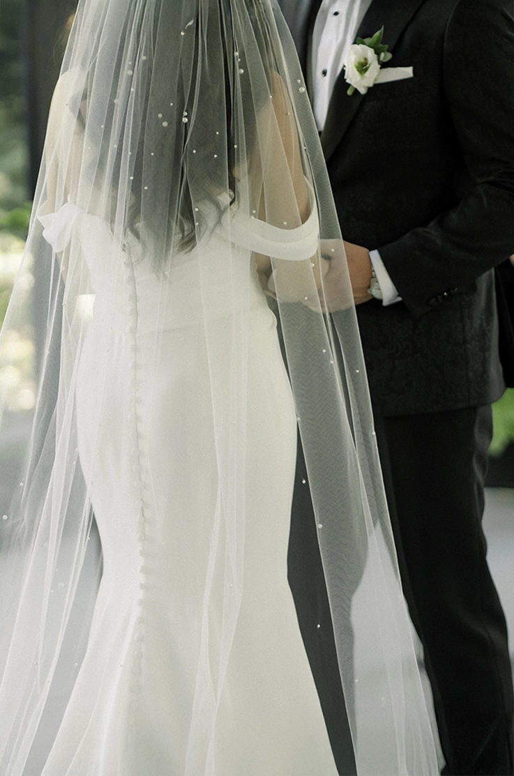 Back of a bride walking in a crepe fit and flare gown with draped sleeves, a buttoned back and pearl veil, facing her groom. 