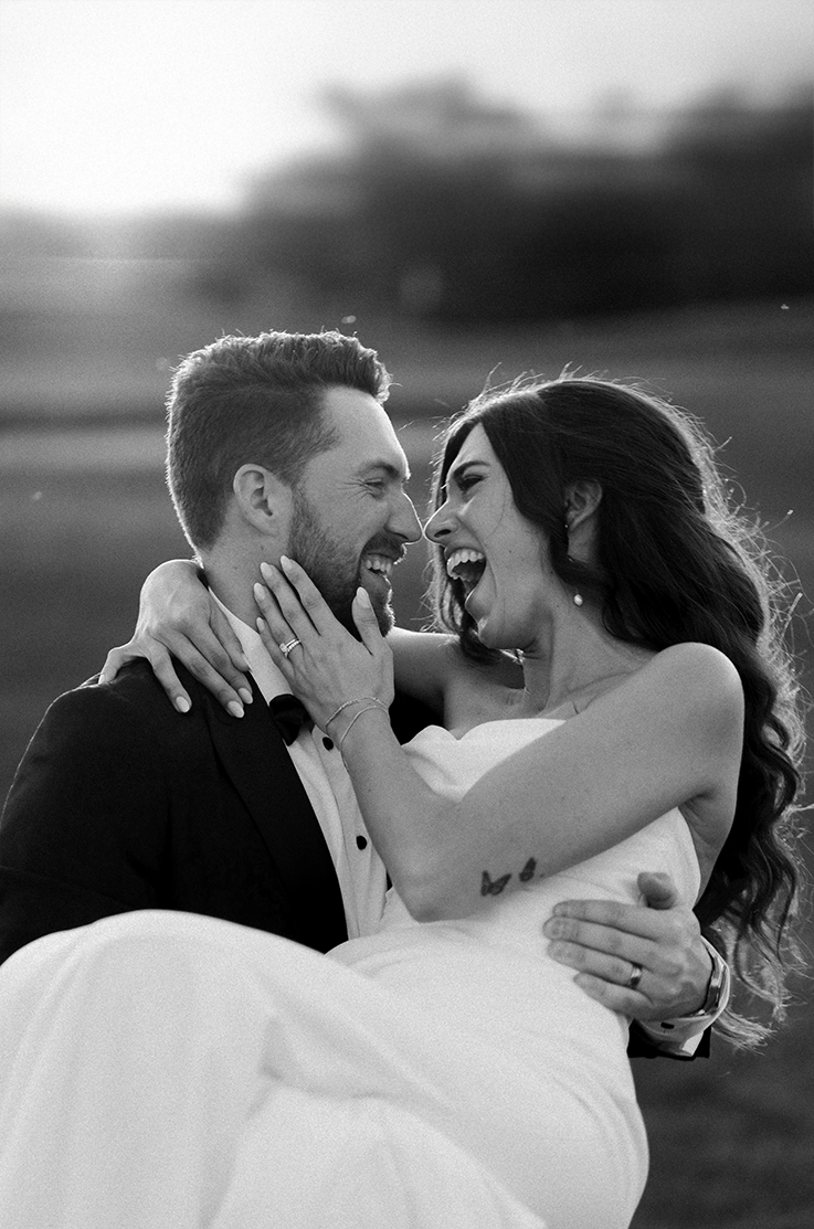 Groom in a tux, picks up his bride in a strapless draped wedding gown in an open field in black and white. 
