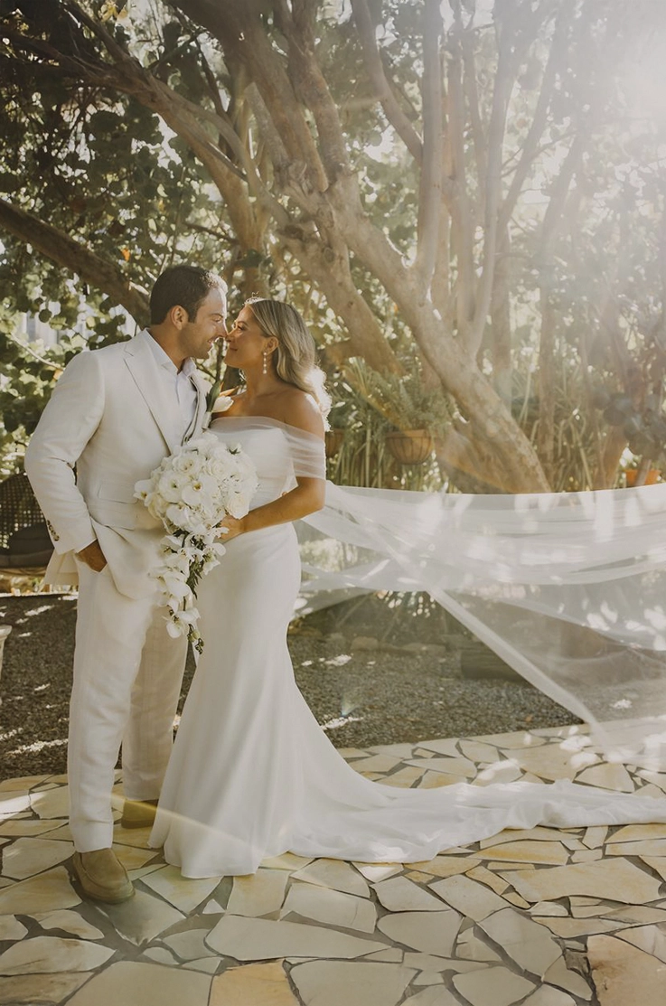 Bride in a draped crepe bodice gown with fit and flare skirt and a flowing veil, posing with groom on an stone path.