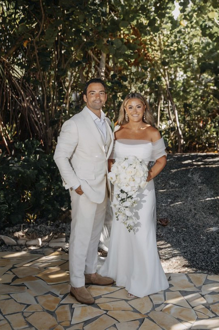 Bride in a crepe fitted wedding dress with draping, holding a large bouquet beside groom in a white suit outdoors.