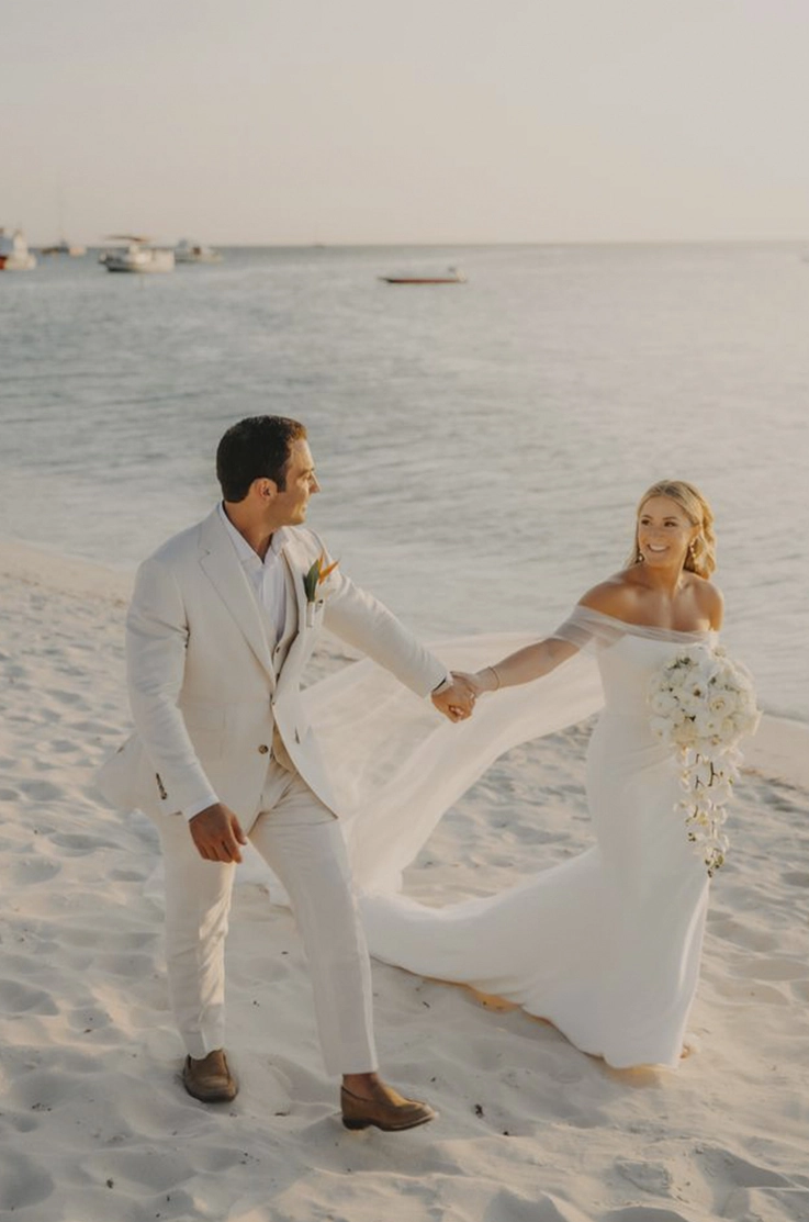 Bride in a crepe fit and flare gown with off-shoulder sleeves, holds a bouquet and walks alongside her groom on a beach.