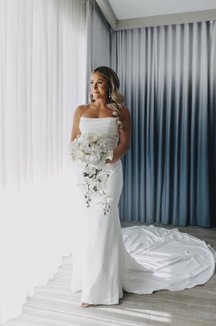 Bride poses in a scoop neck crepe gown with a draped bodice, by a bright window with white curtains in bridal suite.