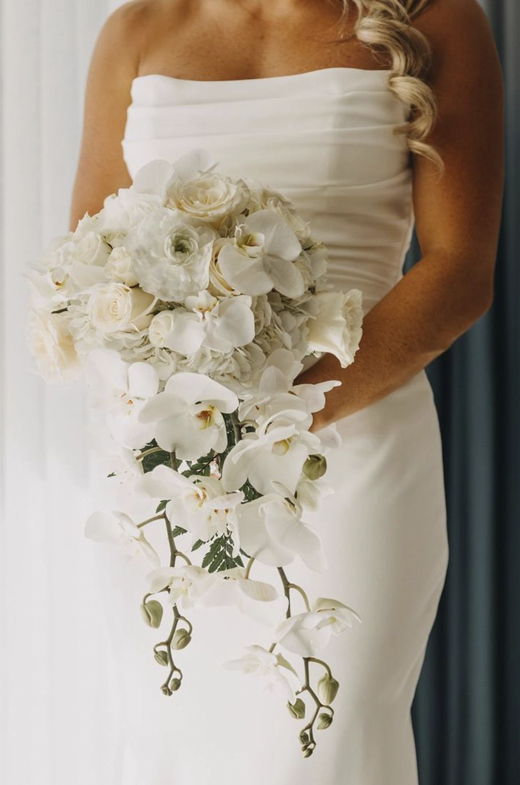 Close-up of bride in a strapless fit and flare gown with a draped bodice, and is holding a bouquet by curtains.