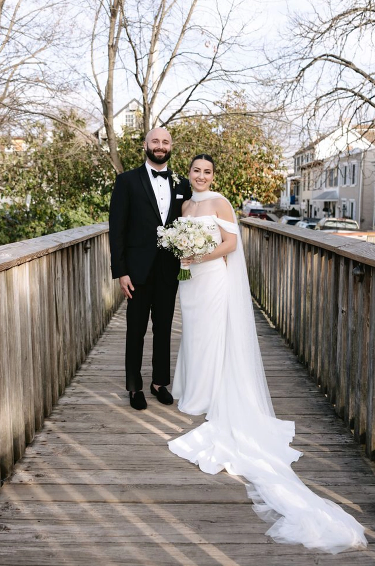 Bride in a scoop neck crepe wedding dress with draping, sleeves and scarf, is posing with groom on a wooden bridge.