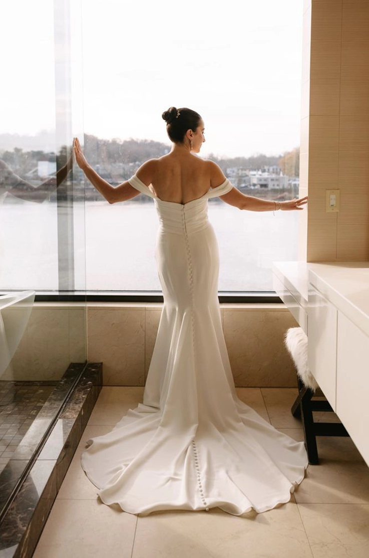 Back of bride in a fit and flare crepe gown with buttons down the back and draped sleeves, posing at a window.