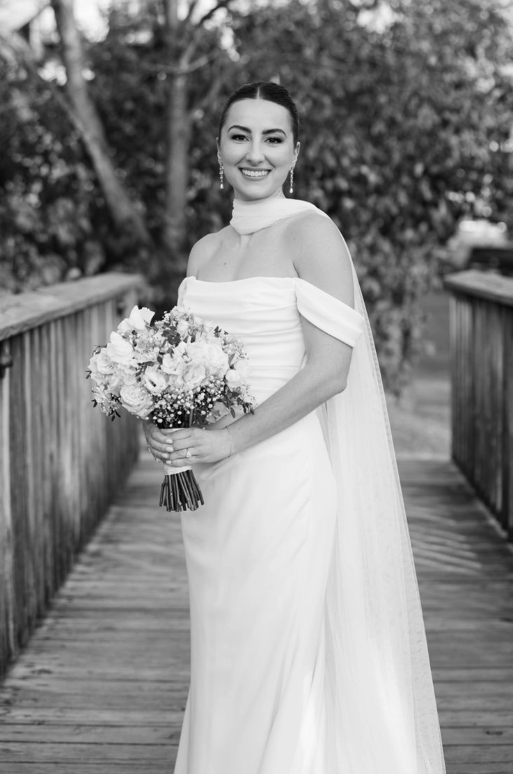  Close-up of bride in a draped scoop neck fit and flare gown with a scarf and off-shoulder sleeves, posing in black and white.