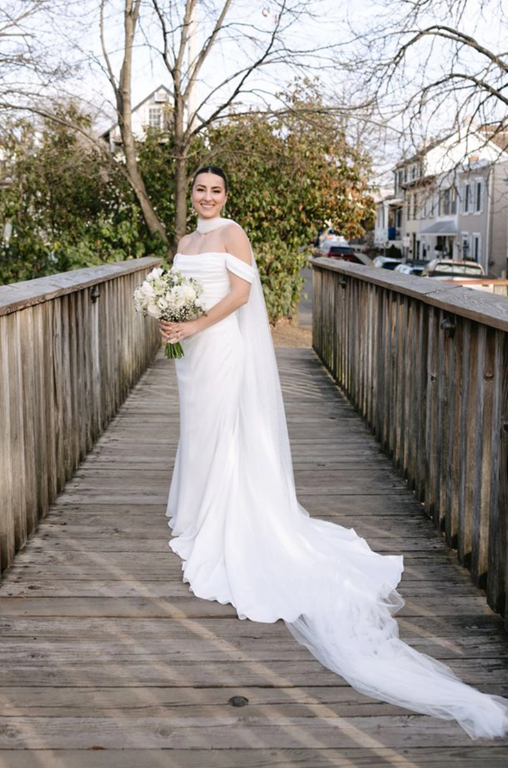 Bride in a draped crepe gown with off-shoulder sleeves, and a flowing scarf, holding a bouquet on a bridge. 