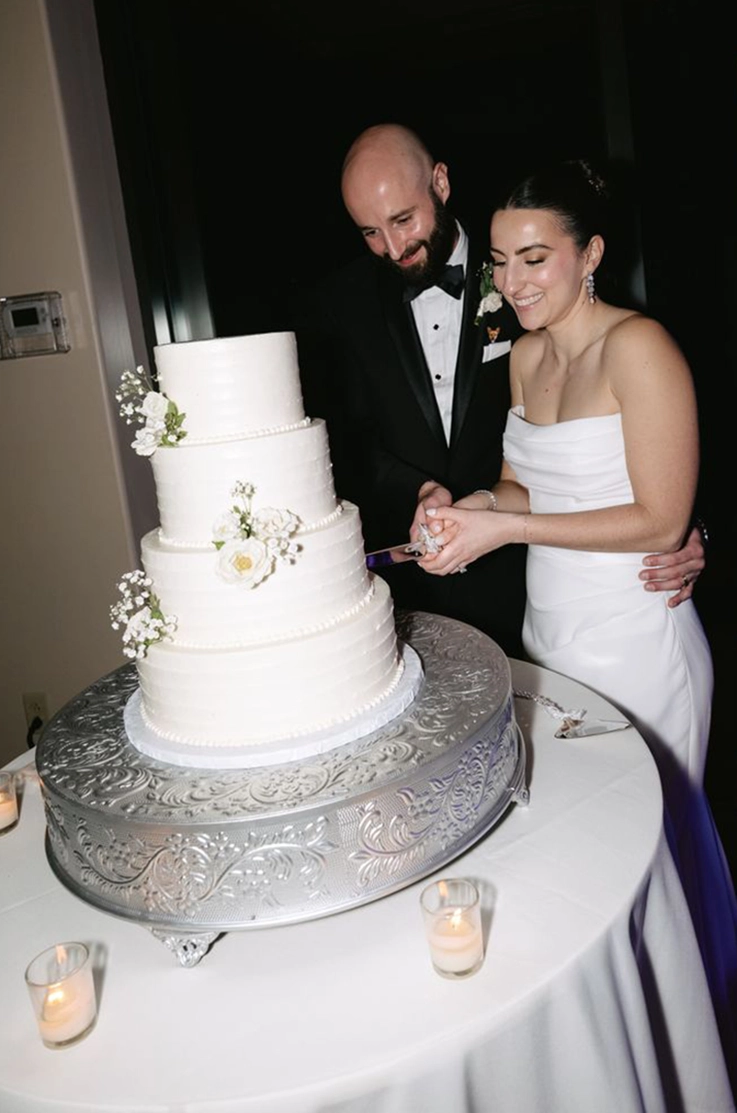 Bride in a crepe fitted gown with a scoop neckline and draping, cuts the cake with her groom in a tuxedo.
