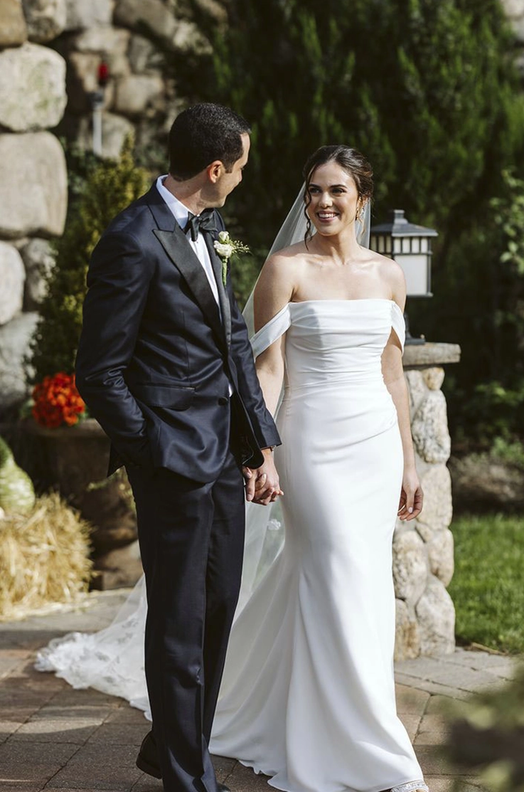Bride in a draped scoop neck gown with fit and flare skirt, sleeves and a veil, holds hands and walks with the groom. 