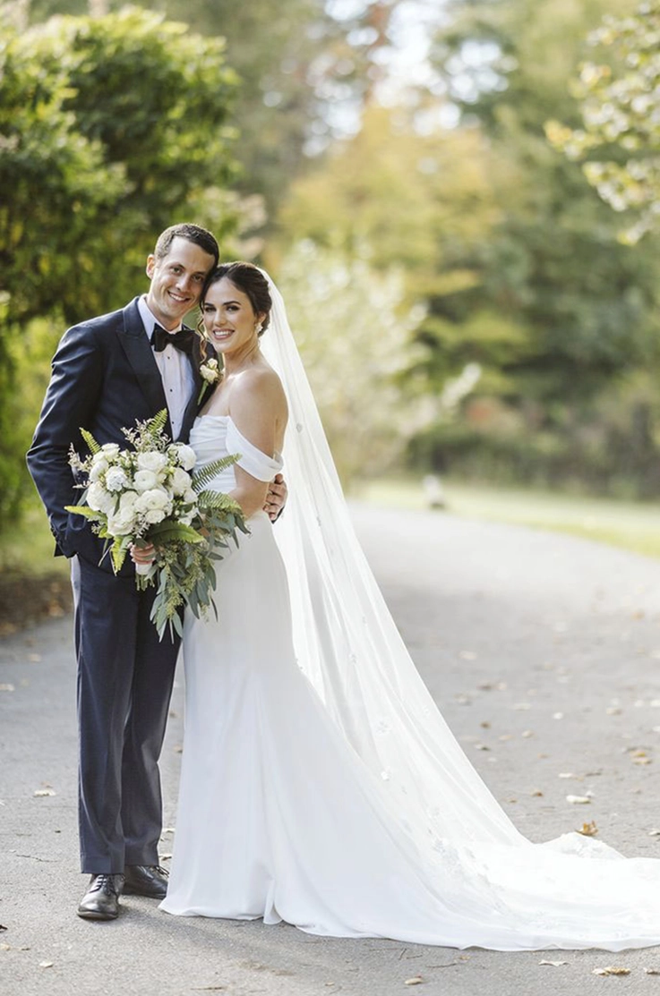 Bride in a crepe fit and flare wedding dress with sleeves, draping and a veil, posing alongside her groom on an open path. 