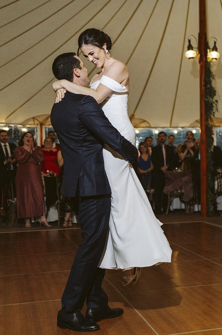 Groom picks up his bride in fit and flare gown with draped sleeves, as they dance in a tented reception.