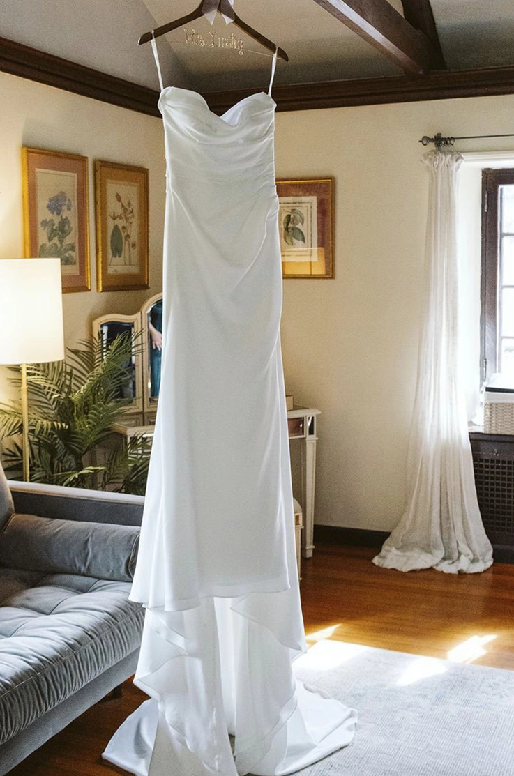 A crepe draped bodice gown with fit and flare skirt hangs from a ceiling in bridal suite by a bright window. 