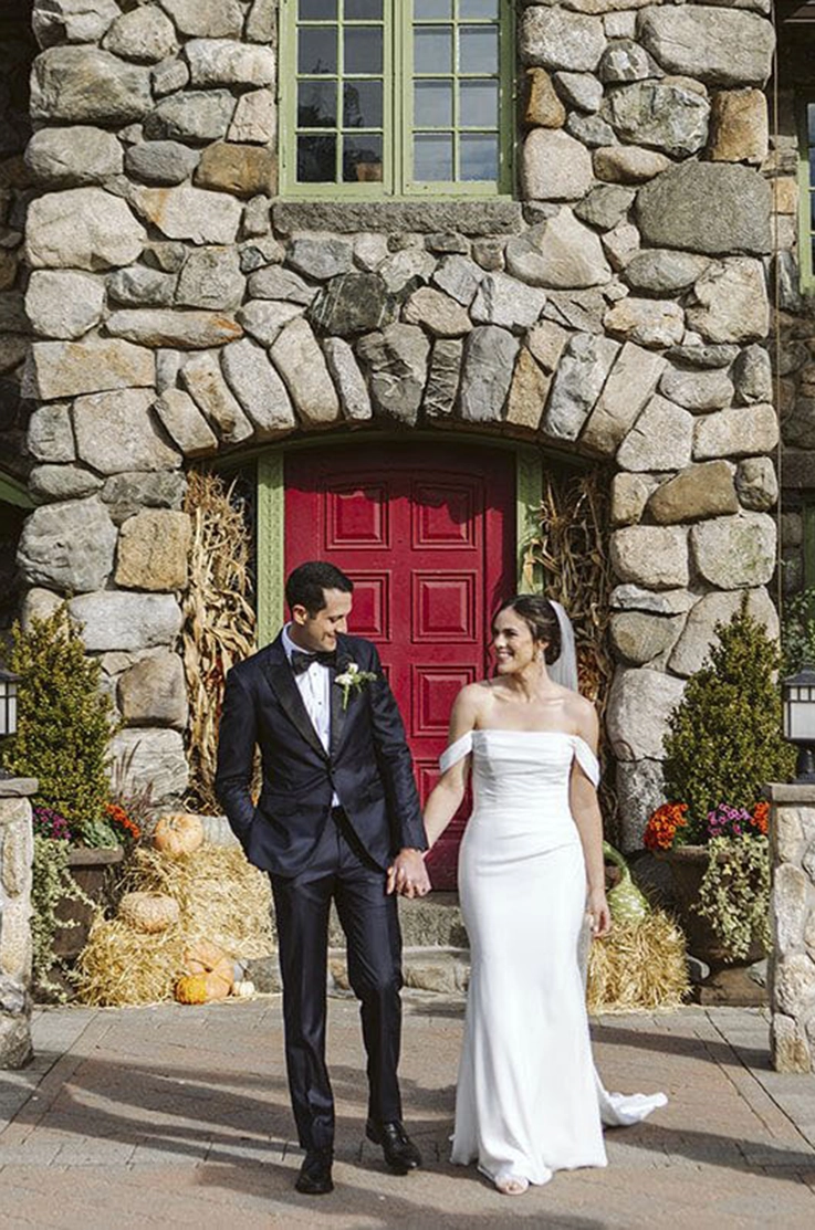 Bride in a scoop neck crepe gown with a draped bodice, sleeves and a veil, walks hand in hand with groom outside the venue.