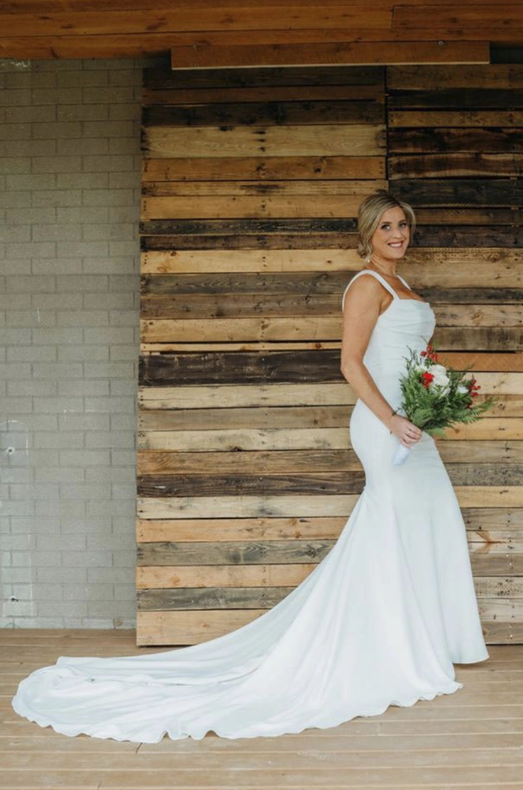 Bride poses in a sleeveless crepe gown with a draped bodice and fit and flare skirt, holding a bouquet indoors.