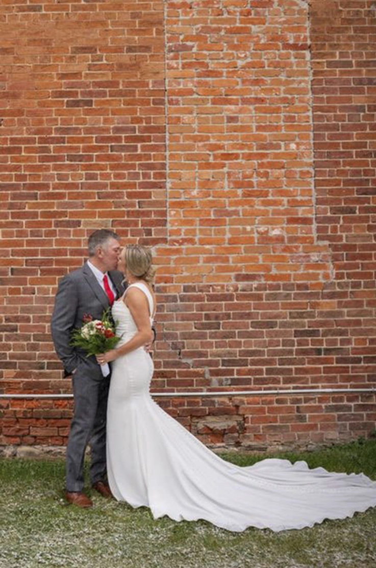 Groom embraces bride in a fit and flare wedding dress with buttons and draping, and they kiss by a red brick wall. 