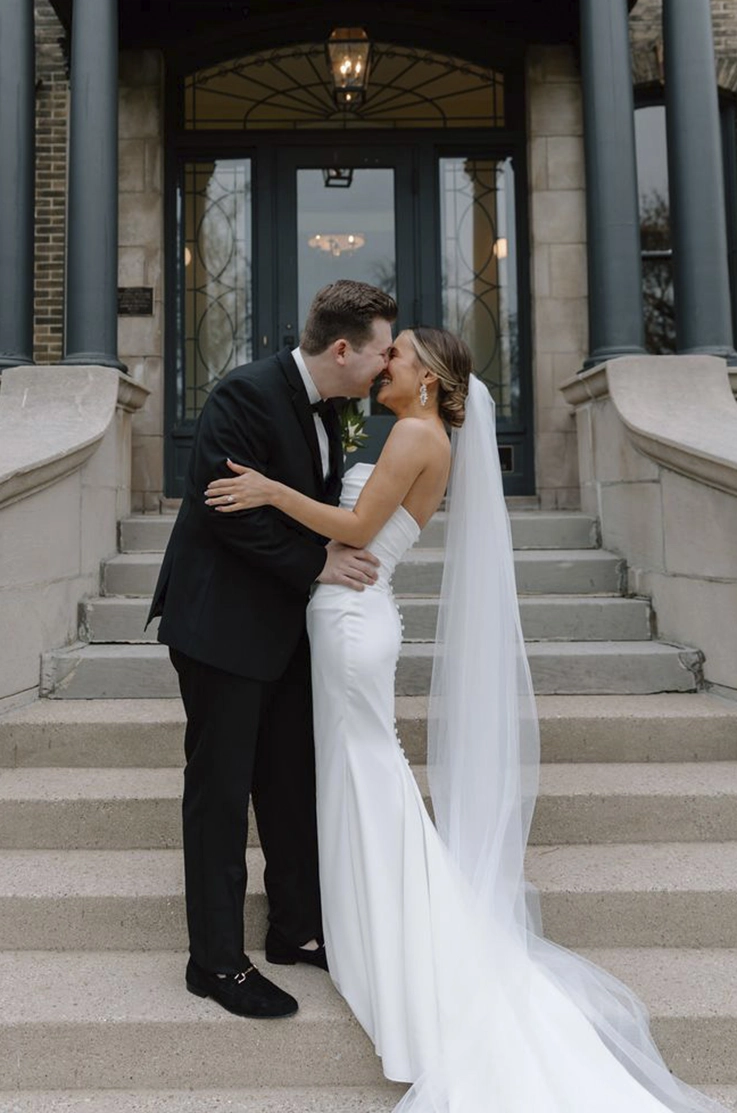Groom kisses his bride in a crepe fit and flare gown with buttons and a veil, and are standing on the stone steps of the venue.