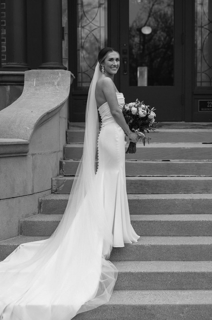 Bride poses in a strapless crepe fitted gown with button detail and a long veil, is holding a bouquet on stone steps.