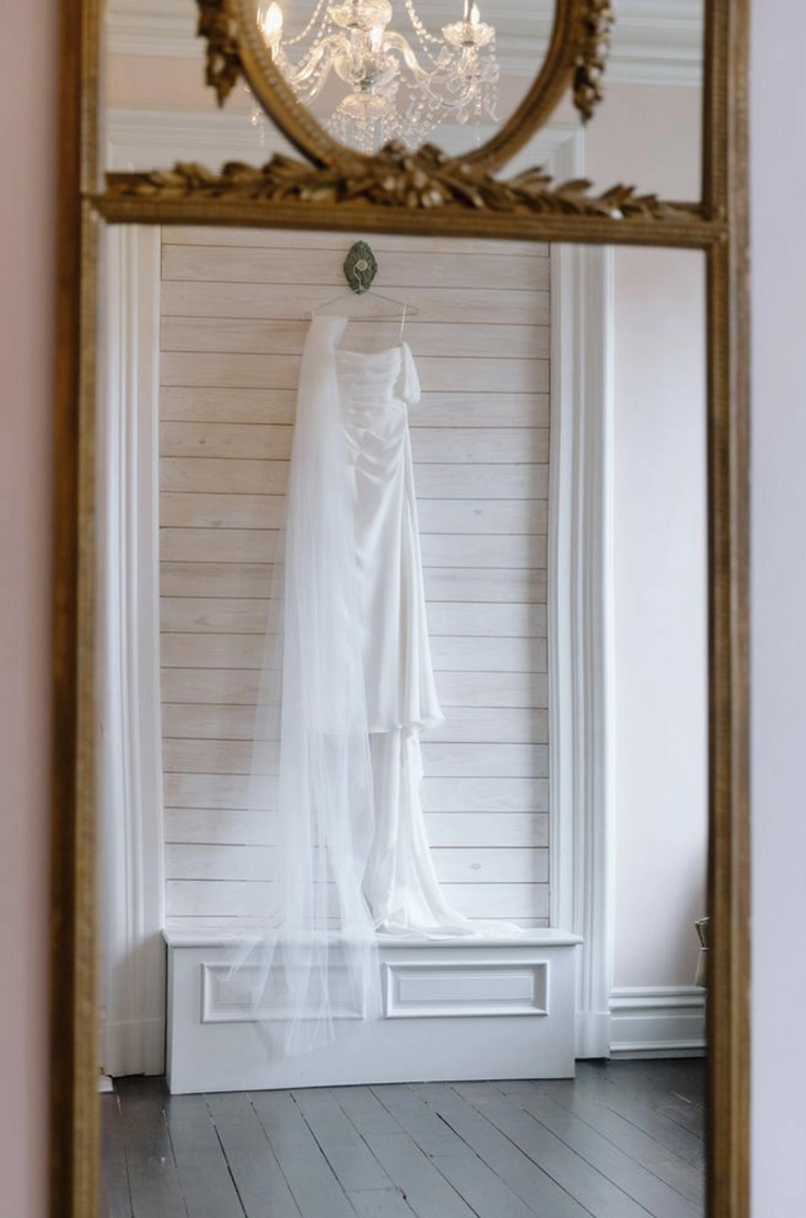 A draped crepe gown with a long veil and off-shoulder sleeves is displayed in a vintage mirror, in a bridal suite. 
