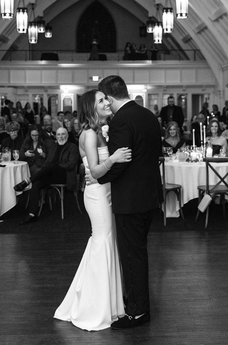 Groom dances with bride in a scoop neck crepe wedding dress with fit and flare skirt, at reception in black and white.
