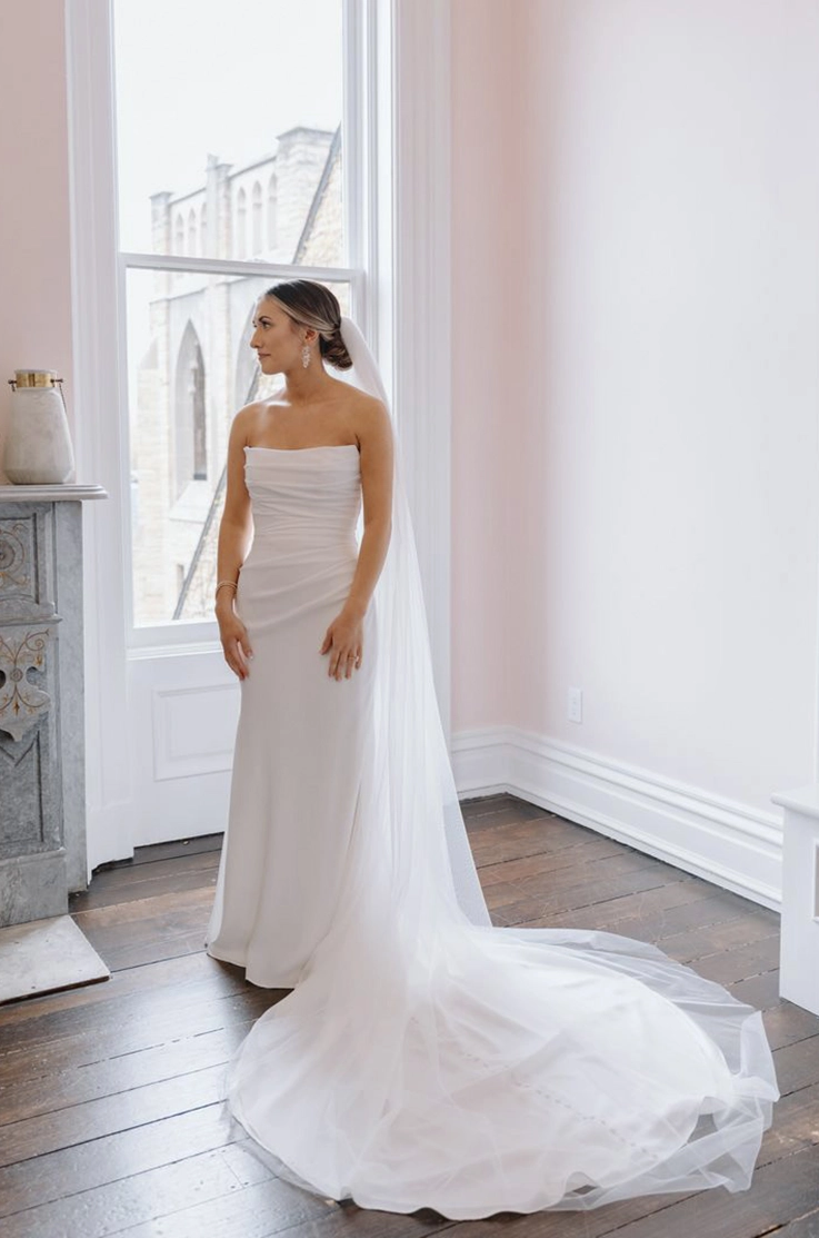 Bride posing in a scoop neck crepe gown with a draped bodice and flowing veil, standing by a window. 