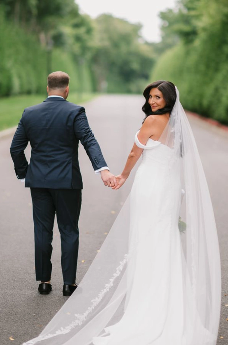 Bride looks over her shoulder in a crepe fit and flare gown with a flowing veil, as she walks hand in hand with her groom. 